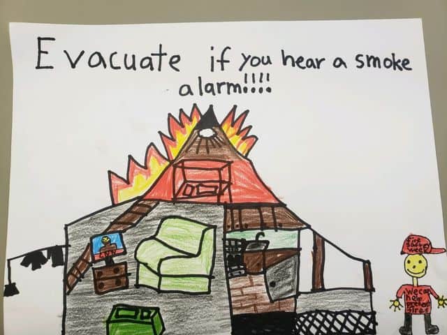 "Evacuate if you hear a smoke alarm" hand drawn image of a house on fire and a person wearing a red hat that says "Fire Safety Week" and a red shirt that says "We can help prevent fires." Art by Matthew B.